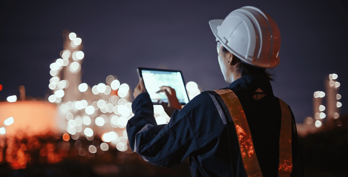 Industrial engineer using a digital tablet at night in front of a brightly lit refinery or industrial plant.