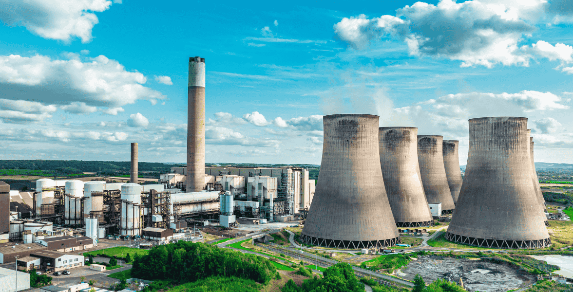 A wide-angle view of a power station with multiple large cooling towers and smokestacks surrounded by green landscape and partly cloudy sky.